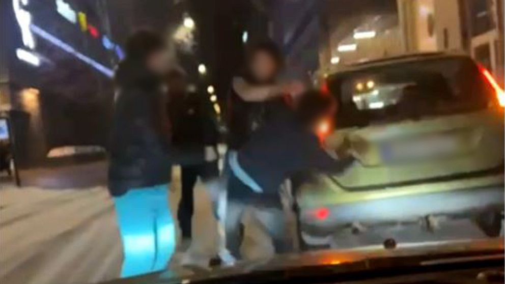 Fyra personer med maskade ansikten syns slåss utanför en bil. En av personerna lyfter sin högra arm mot en av de andra.