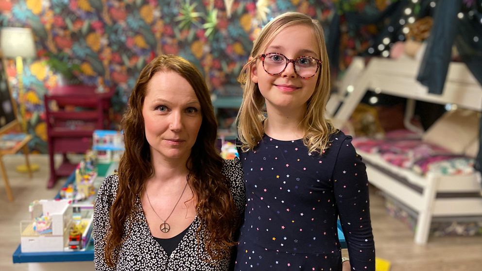 En kvinna och en liten flicka står tillsammans i ett färgglatt rum med mönstrad tapet och leksaker på golvet. Rummet har en familjär atmosfär med en säng och en stol i bakgrunden.