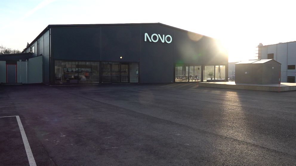 Industrihall med skylten NOVO, tom parkeringsyta och motljus från solnedgång.