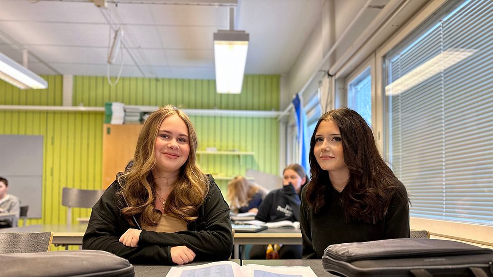 Två tjejer sitter i en klassrumsmiljö och har ett häfte uppslaget framför sig.