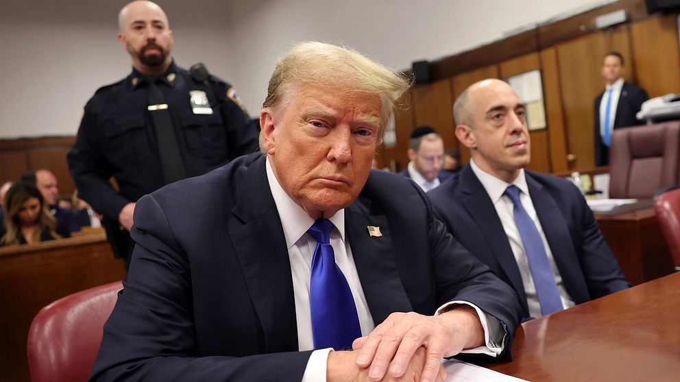 Donald Trump sitter vid ett bord i en rättssal. En av dem verkar vara en juridisk representant, medan ytterligare en person i uniform står i bakgrunden. Rättssalen har mörka träväggar och en formell atmosfär, med stolar och ett dokument på bordet framför dem.