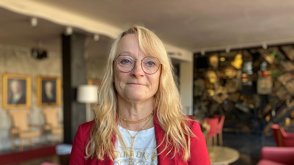 En person står framför en modern interiör med en vägg av målningar i bakgrunden. Personen bär en röd kavaj och en vit t-shirt med text. Rummet har en stilren möblering med rosa soffor och ett runt bord.