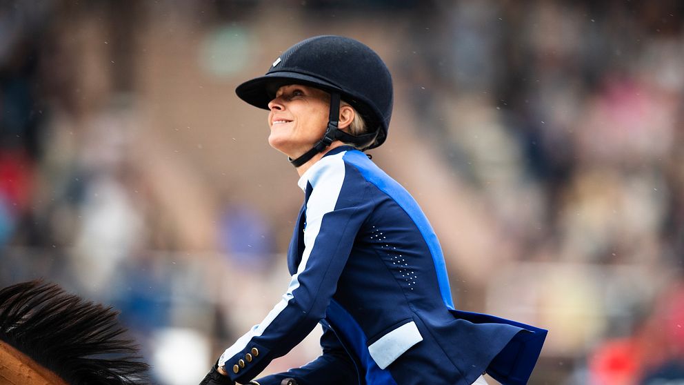 Ryttare i blå och vit tävlingsdräkt på en häst, fångad i en dynamisk pose under en tävling. I bakgrunden syns en oskarp publik som bidrar till den sportiga atmosfären.