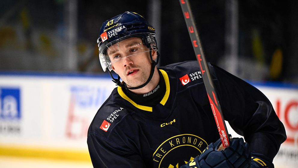 En ishockeyspelare från Tre Kronor i mörkblå utrustning och hjälm, håller en röd ishockeyklubba vid sidan av isen med reklamskyltar i bakgrunden.
