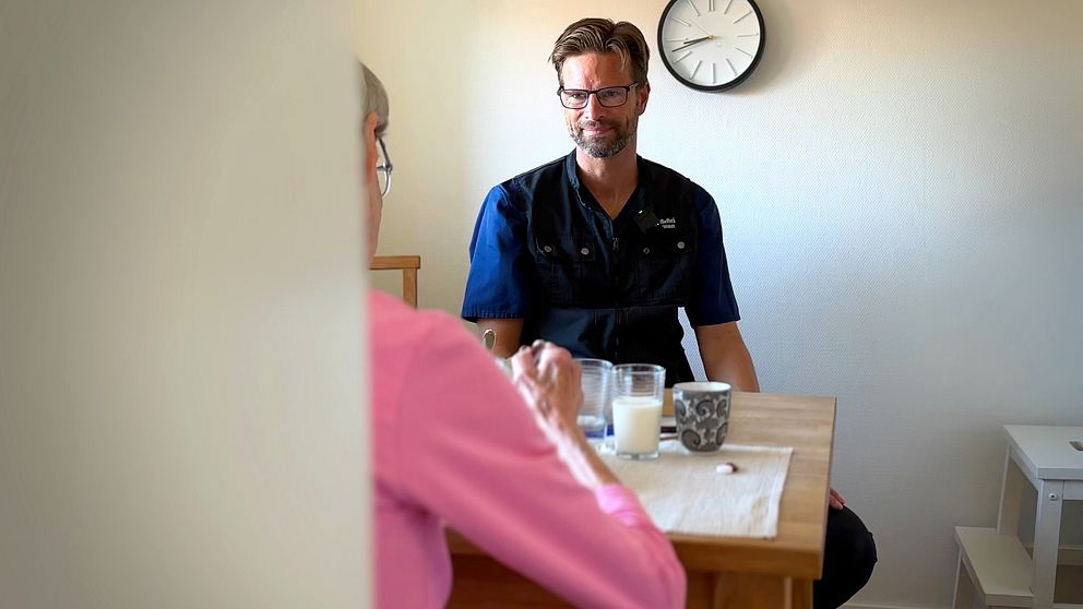 Mathias Rönngren i hemtjänst sitter med kvinna i rosa tröja vid matbord