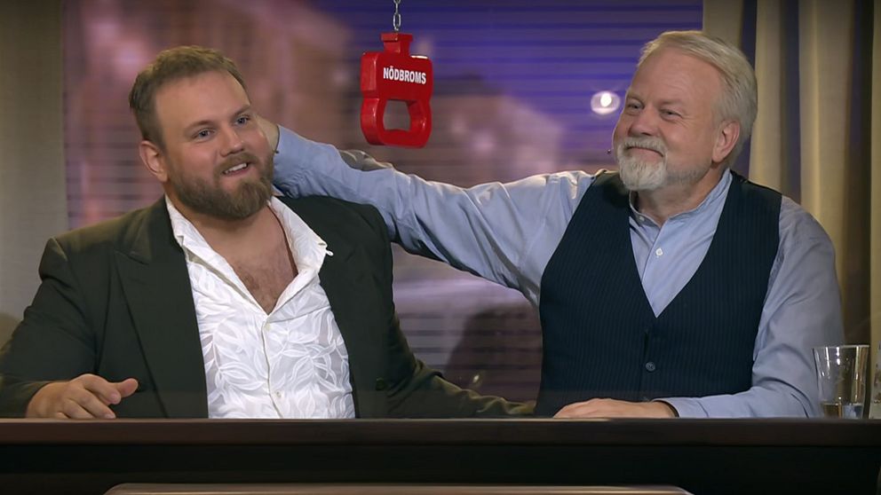 Två män sitter vid ett bord i en studiomiljö med en mikrofon hängande mellan dem. En av männen har sin arm över den andres axlar.
