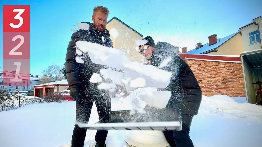 Två personer står i snön, den ena skyfflar snö mot kameran.
