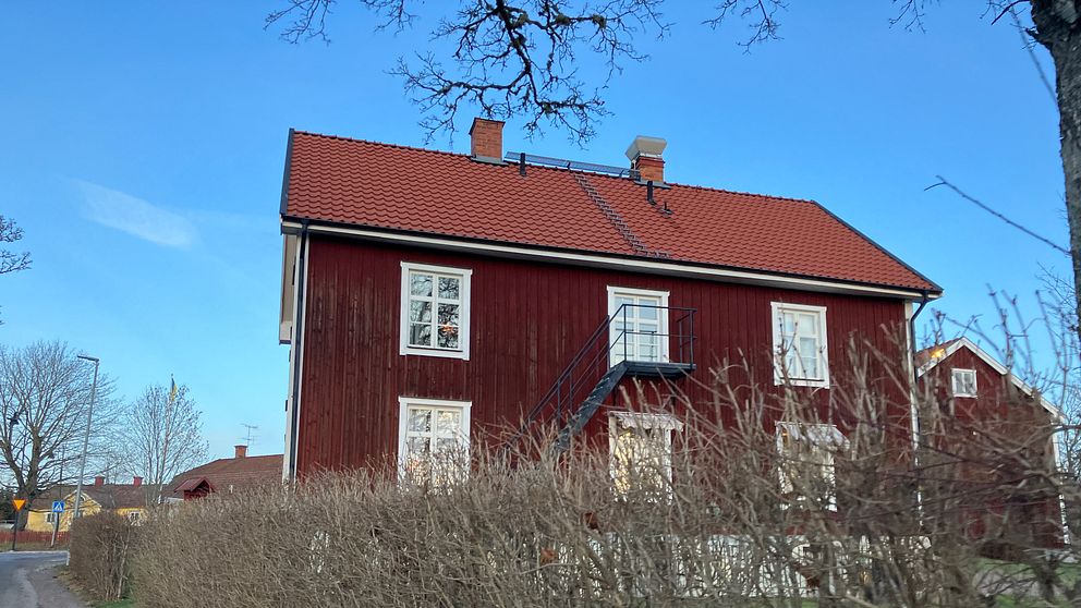 Ett rödmålat hus med vitmålade fönster och en balkong, omgiven av en tät häck och beläget vid en grusväg. Himlen är klar blå och det finns träd i bakgrunden.