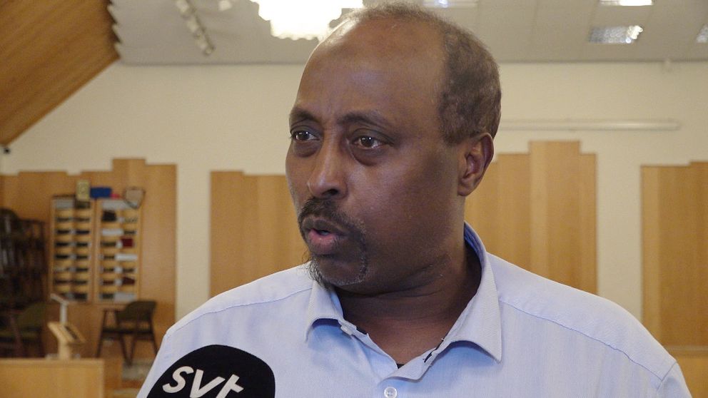En person står i ett rum med träpaneler på väggarna. I förgrunden hålls en mikrofon märkt med ”SVT”, vilket tyder på en intervjusituation i en studiomiljö eller ett offentligt uttalande.