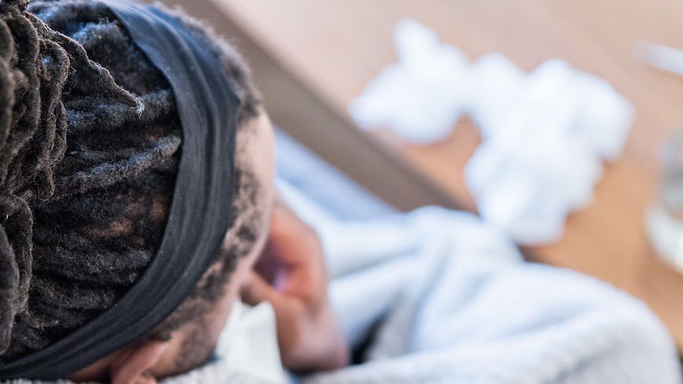 En person sitter svept i en filt och ser eftertänksam ut, med en hand som stöder hakan. I bakgrunden syns en hög med använda näsdukar på bordet.