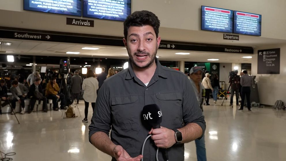 Fouad Youcefirapporterar i en flygplatshall. I bakgrunden syns andra nyhetsteam med kameror och lampor. På väggarna syns tv-skärmar med ett informationsmeddelande och skyltar där står Arrivals och Departures.