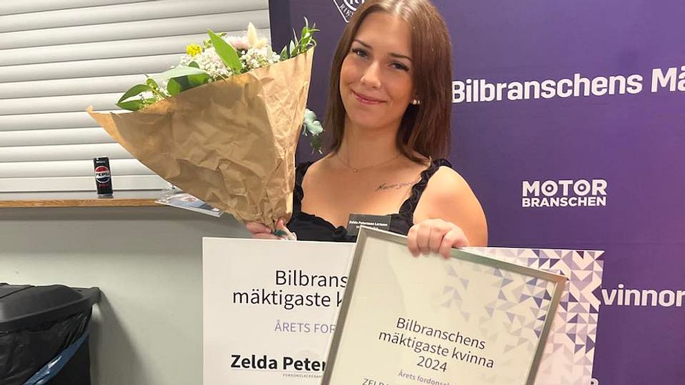 Zelda Pettersson Larsson i en svart klänning håller en bukett blommor i ena handen och ett diplom i den andra. Diplomet visar att personen har vunnit priset ”Bilbranschens mäktigaste kvinna 2024”. Bakgrunden innehåller en banner med texten ”Bilbranschens mäktigaste kvinna” samt logotypen för Motorbranschen.