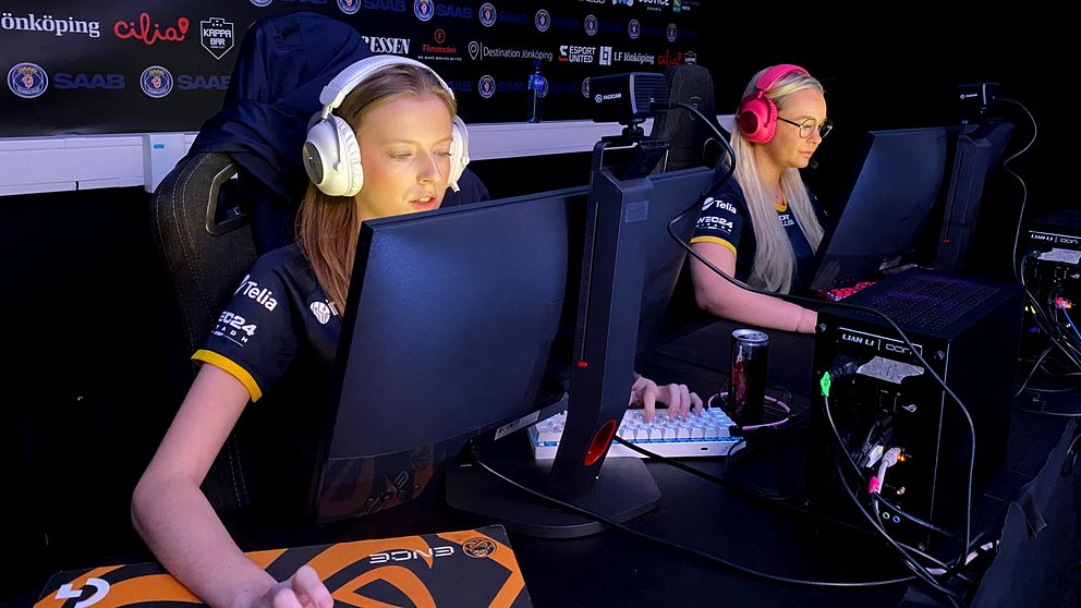 Två kvinnliga gamers sitter vid sina datorer under en e-sporttävling. Landslagspelarna Emma Mattsson och Linnéa Claesson. Den första spelaren har på sig hörlurar och är fokuserad på skärmen, medan den andra också bär hörlurar och använder ett tangentbord. Båda spelar i en mörk miljö med lagets logotyper synliga i bakgrunden.