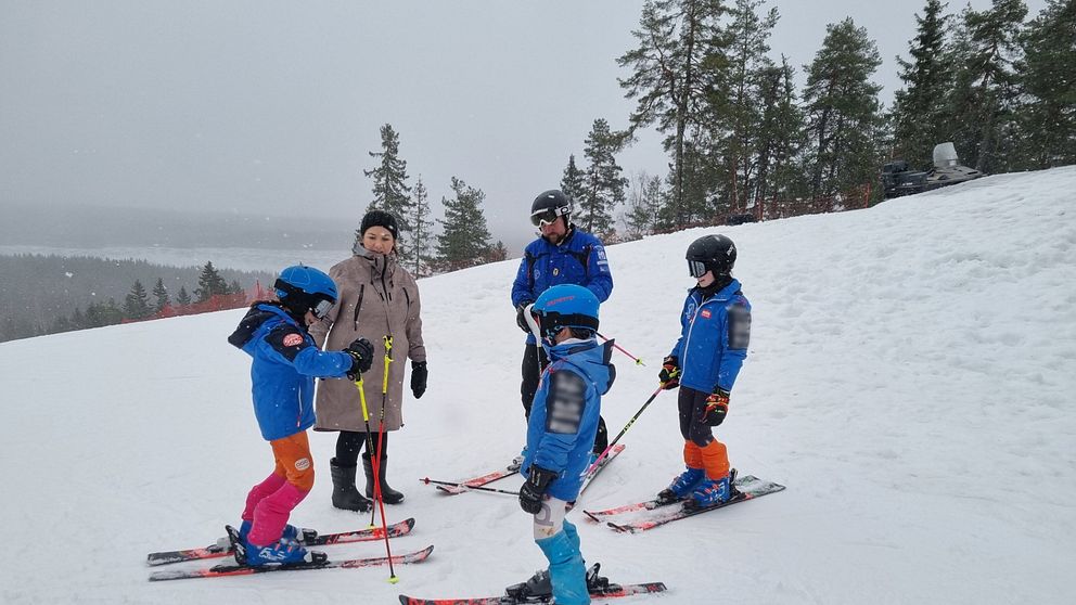 En grupp barn i skidkläder står på snön vid en skidbacke. Två av barnen är upptagna med att justera sina skidor medan den vuxna och andra barn tittar på. Det snöar lätt och det syns träd i bakgrunden.