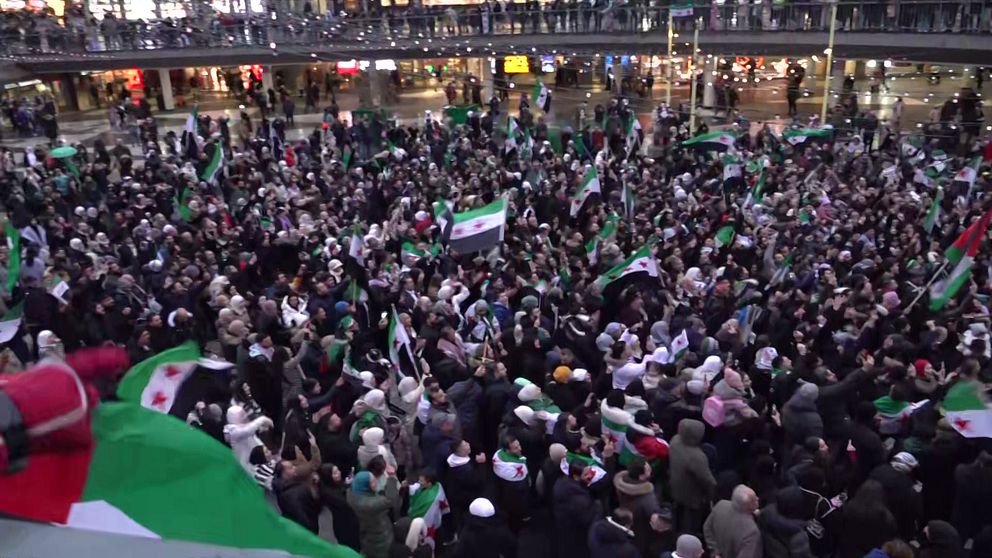 Stort folkmassa som håller upp syriska flaggor under en demonstration. Människorna är samlade på en offentlig plats, klädda i varierande kläder, och det är en känsla av gemenskap och engagemang. Bakgrunden visar en livlig stadsbild med belysning och byggnader.