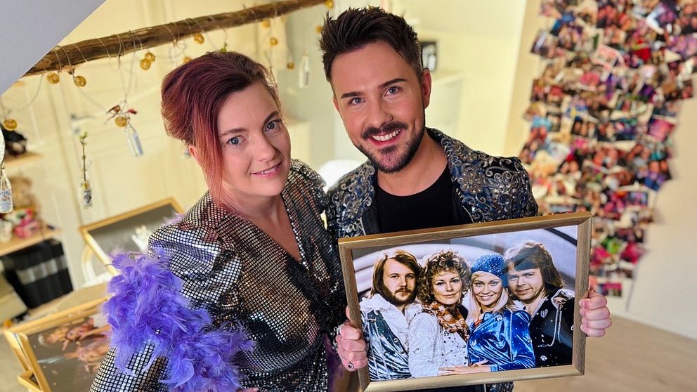 Två personer klädda i glittriga kläder håller i en inramad fotografi med Abba. De står i ett utrymme dekorerat med en vajer fylld med bilder och färgglada klädnypor i bakgrunden.