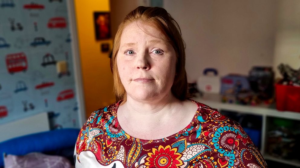 Cecilia, förälder som har svårt att få ekonomin att gå ihop, står i ett rum klädd i en färgglad blommig blus. I bakgrunden syns väggar dekorerade med bilder av bilar. På en hylla i bakgrunden finns förvaringslådor och leksaker.