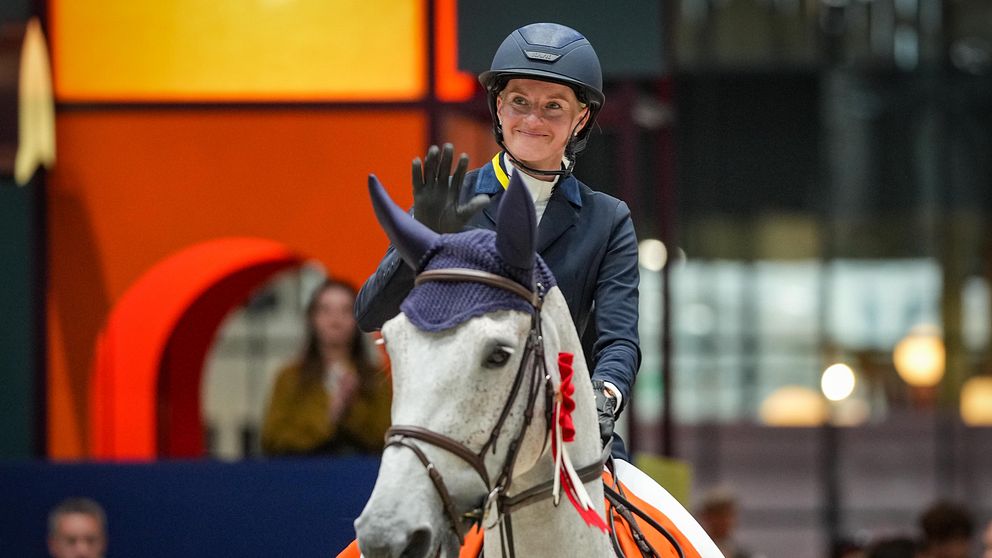 En ryttare i en mörk ridsportjacka och hjälm, sitter på en vit häst som är utrustad med en blå sadel och en orange täcke. Ryttaren viftar med hand en uttryck av seger eller uppskattning, omgivet av en färgglad bakgrund med geometriska former.