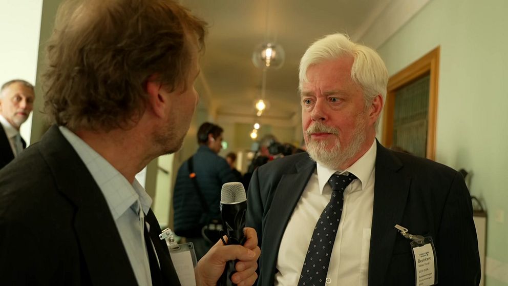 Reporter intervjuar Niklas Ekvall i kostym i en korridor, flera personer syns i bakgrunden.