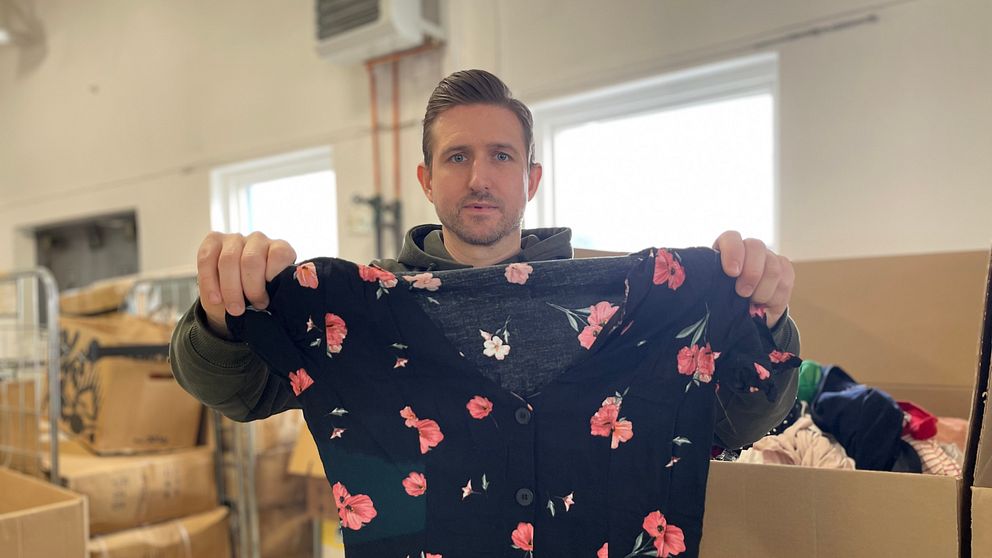 En person håller upp en svart klänning med röda blommor i vad som verkar vara ett lager- eller secondhand-miljö, omgivet av stora kartonger och lagerhyllor.