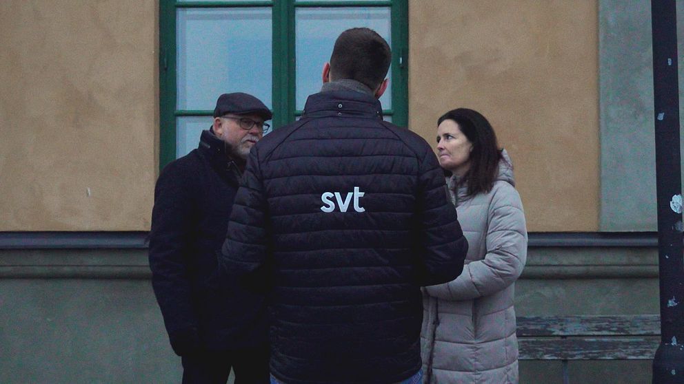 En reporter från SVT intervjuar två personer utomhus vid en byggnad med grön fönsterlucka. Reportern bär en svart jacka med SVT-logotyp på ryggen.