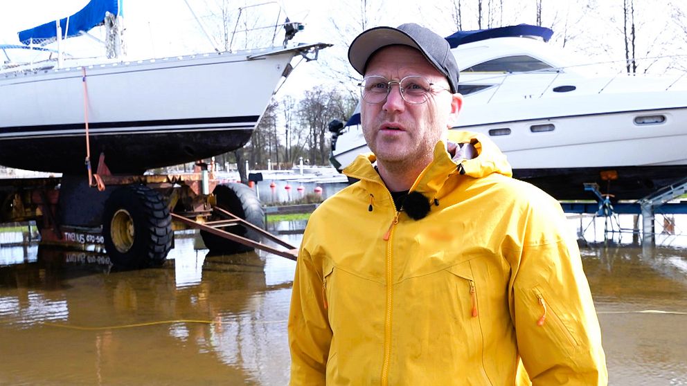 Man i gul jacka med svart keps. Står på land framför båt. I bakgrunden syns översvämmat mark.