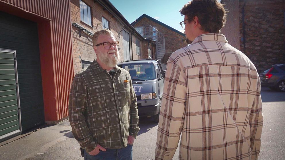 Två personer samtalar utomhus vid en bakgata med tegelbyggnader och en parkerad bil.
