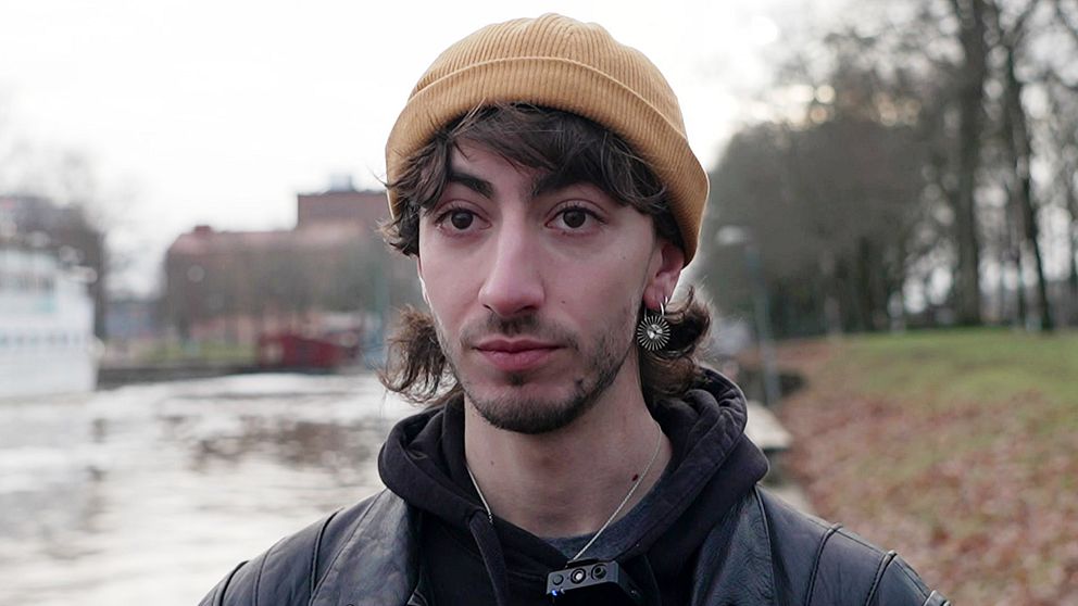 Louka Bertolino har en senapsgul mössa på sig och örhänge. Han har mörka ögon och skäggstubb. Han har på sig en svart skinnjacka och en svart hoodie under den.