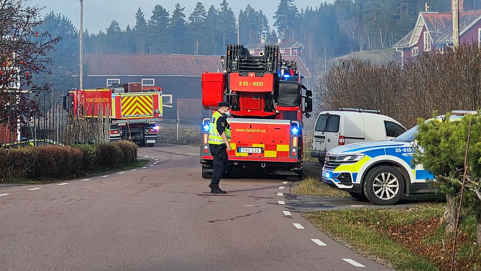 Brandbilar och en polisbil på en väg i Rättvik. En polisman med skydd för ansiktet står på vägen och rök syns i luften.