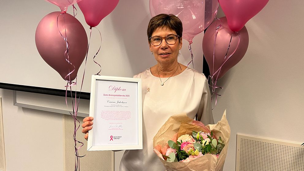En kvinna står vid rosa ballonger och håller upp ett diplom.