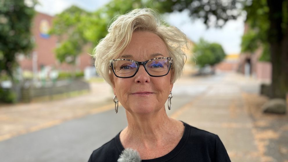 Kvinna med mikrofon står utomhus på en gata med träd och byggnader i bakgrunden.