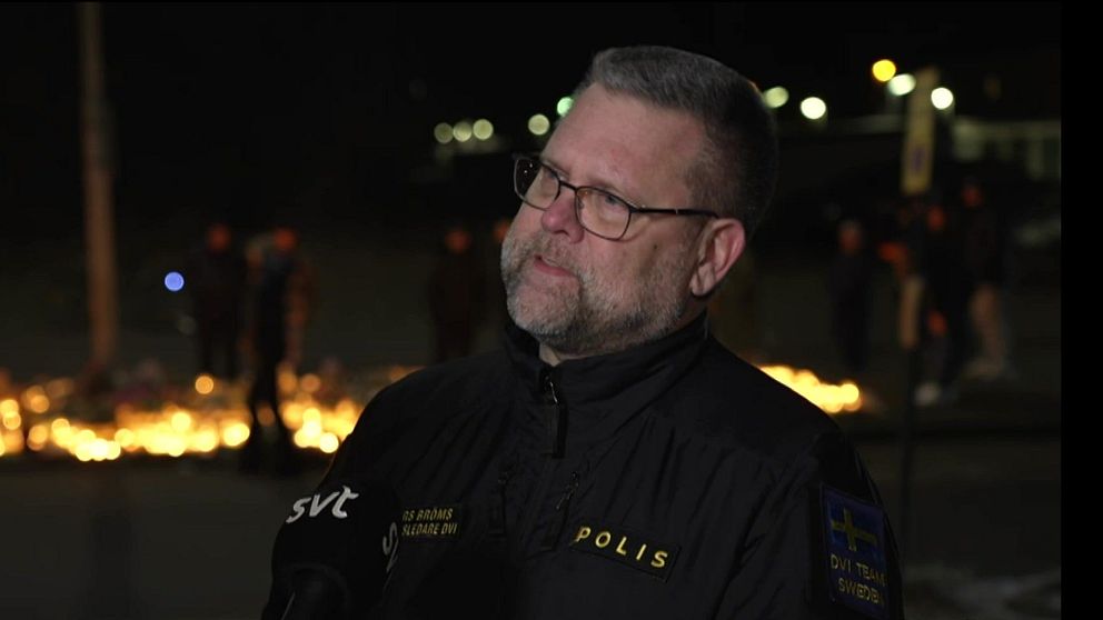 En polis står framför en minnesplats med tända gravljus på en mörk plats, med en mikrofon från SVT i närheten. Olika personer syns i bakgrunden.