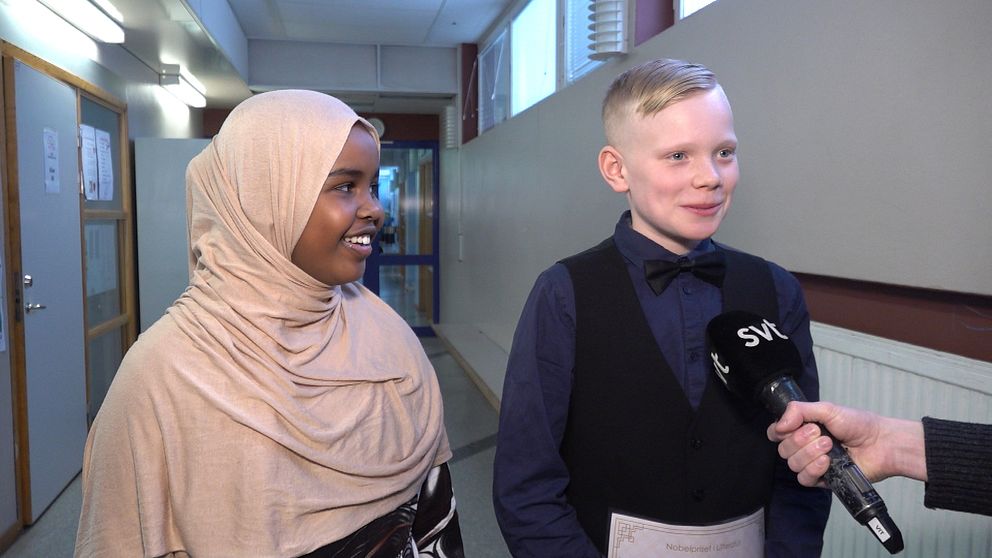 Två barn, en pojke och en flicka, står finklädda och ler i en skolkorridor. Framför sig har de en mikrofon som det står SVT på.