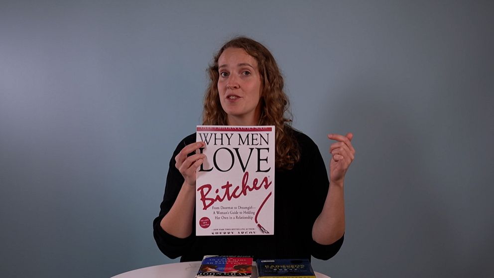 En person håller upp boken ”Why Men Love Bitches” framför sig, med en svart tröja på och en hand gestikulerande i luften. På bordet framför personen ligger två andra böcker synliga. Bakgrunden är ljusblå.
