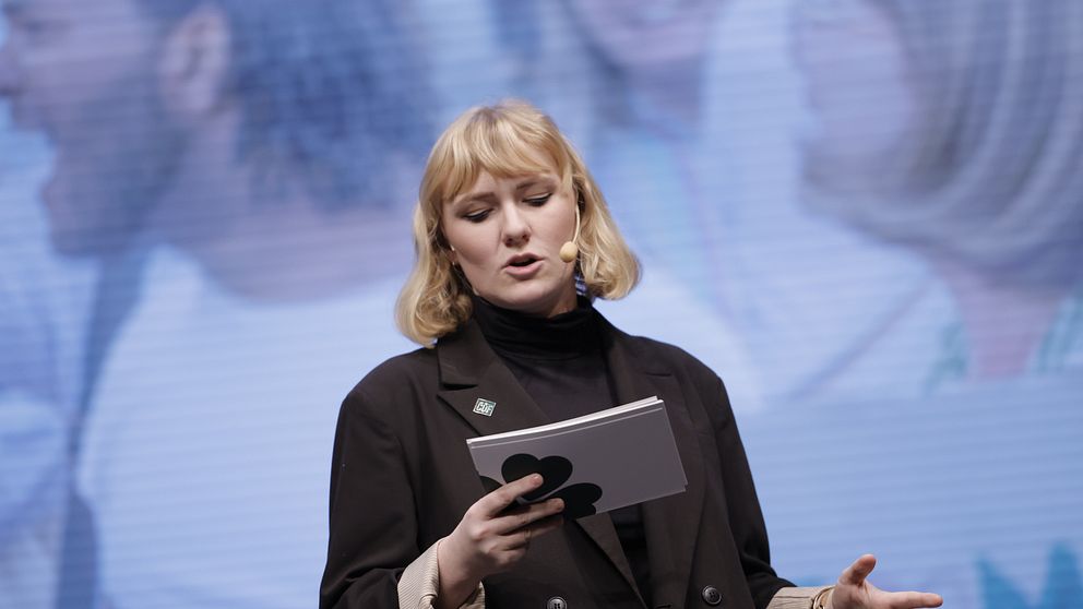 Caroline von Seth talar framför en suddig bakgrund under ett evenemang eller en konferens, med en mikrofon i handen och ett block eller anteckningar i den andra handen.