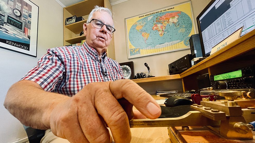 Man sänder morsekod vid ett skrivbord med radioutrustning, karta och dator i bakgrunden.