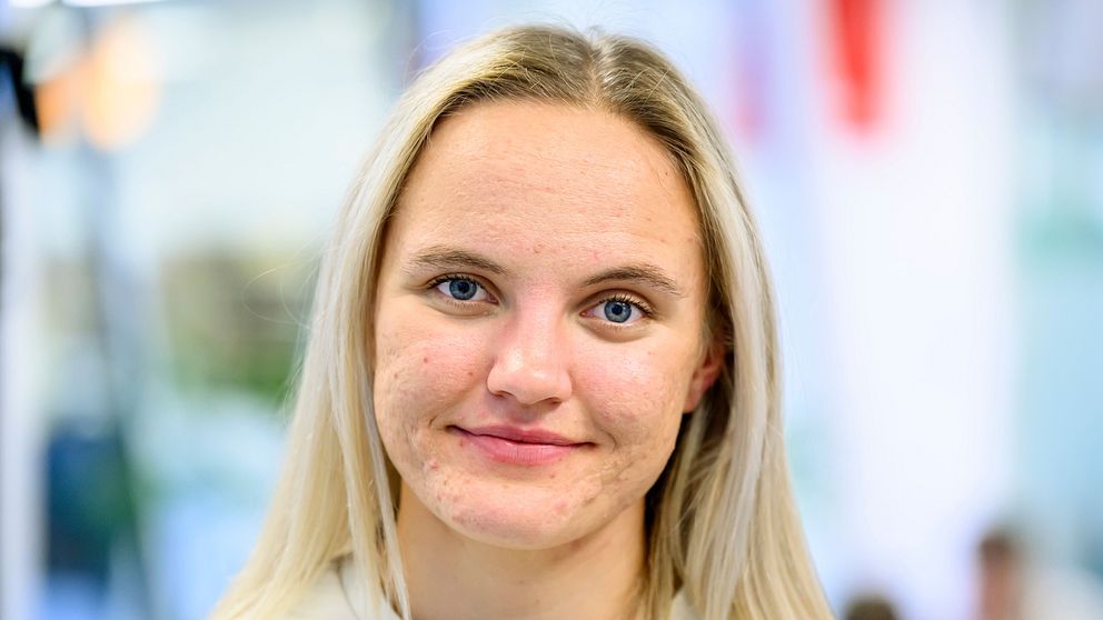 Linn Svahn med långt blont hår står i en inomhusmiljö. Bakgrunden är suddig, med otydliga former och färger som indikerar en social eller professionell sammankomst.