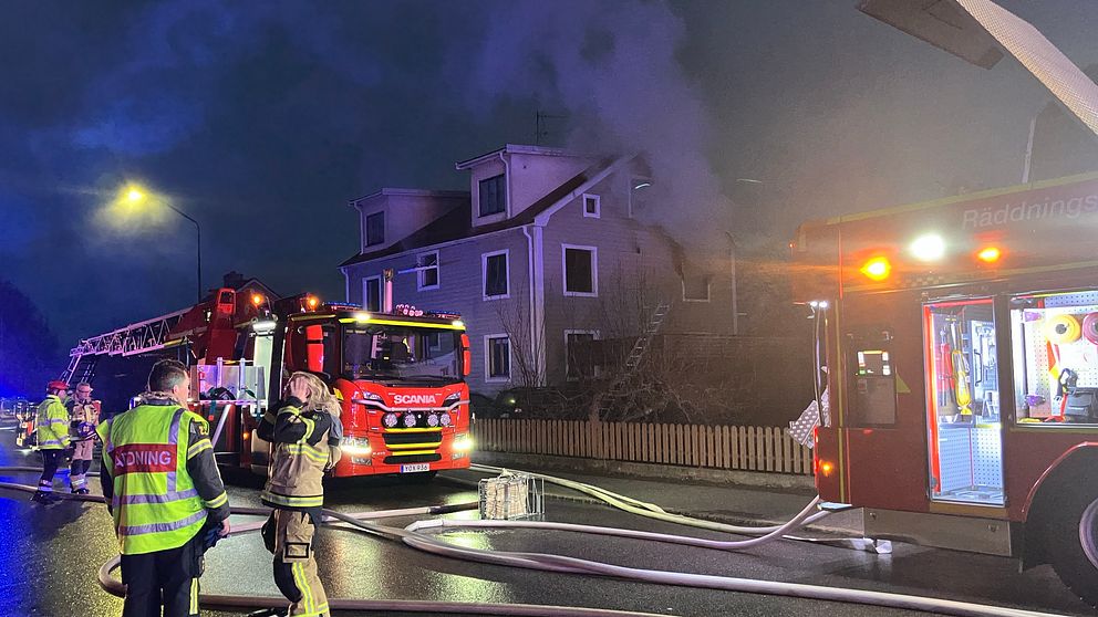 Rök kommer från fönster på andra våningen i tvåvåningshus. Mörkt på platsen med gatlyktor. Räddningstjänst med två bilar och personal på vägen utanför huset.