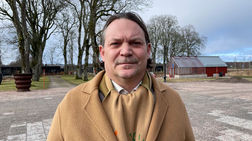 Person i beige kappa står utomhus med träd och byggnader i bakgrunden.