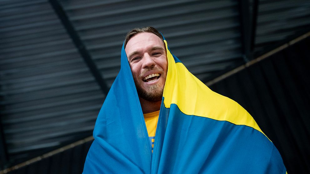 Person inlindad i svensk flagga under ett tak med metallpaneler.