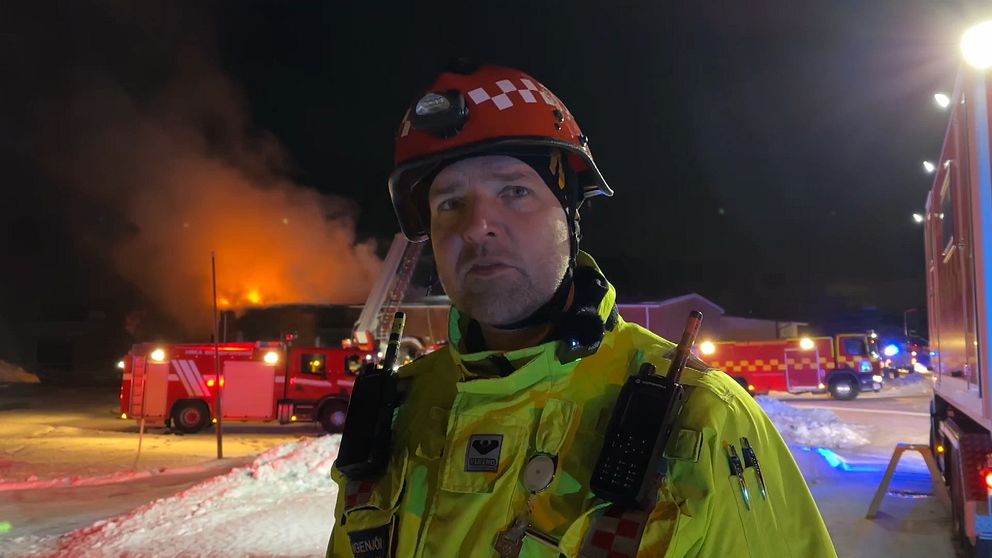 En brandman i röd hjälm och gul uniform står framför en brinnande byggnad och flera räddningsfordon.