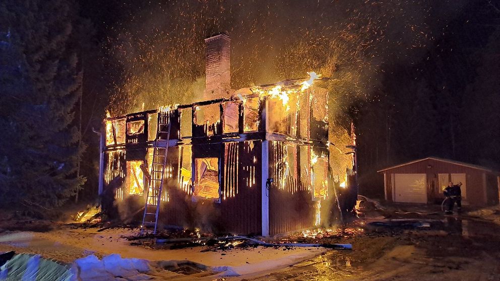 Ett hus som brinner kraftigt med lågor och gnistor som sprakar ut från taket och väggarna, en stege lutad mot fasaden, medan snön på marken reflekterar ljuset från elden.