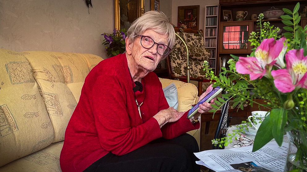 87-åriga Kirsten sitter i en soffa i sitt hem och håller i en mobiltelefon.