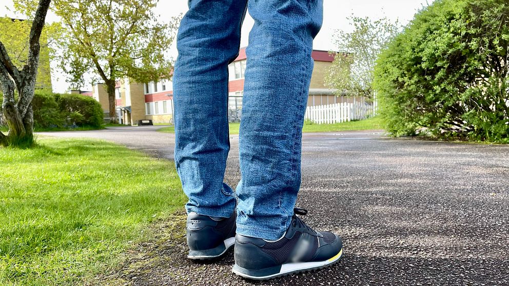 Person i jeans och sneakers står på en asfalterad gångväg i grönskande parkmiljö.