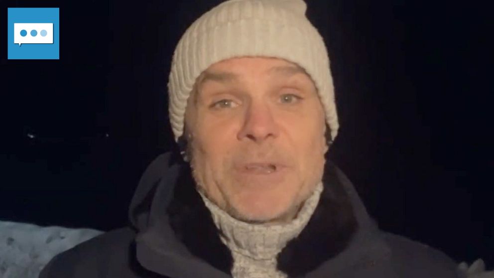 Personen är klädd i en mörk jacka och en ljus mössa, stående utomhus i en mörk miljö. Snö är synlig i bakgrunden, vilket indikerar vinterförhållanden.