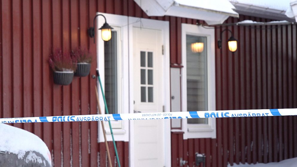 Ett rött trähus med snö på marken och blåvit polisavspärrning framför ingången.