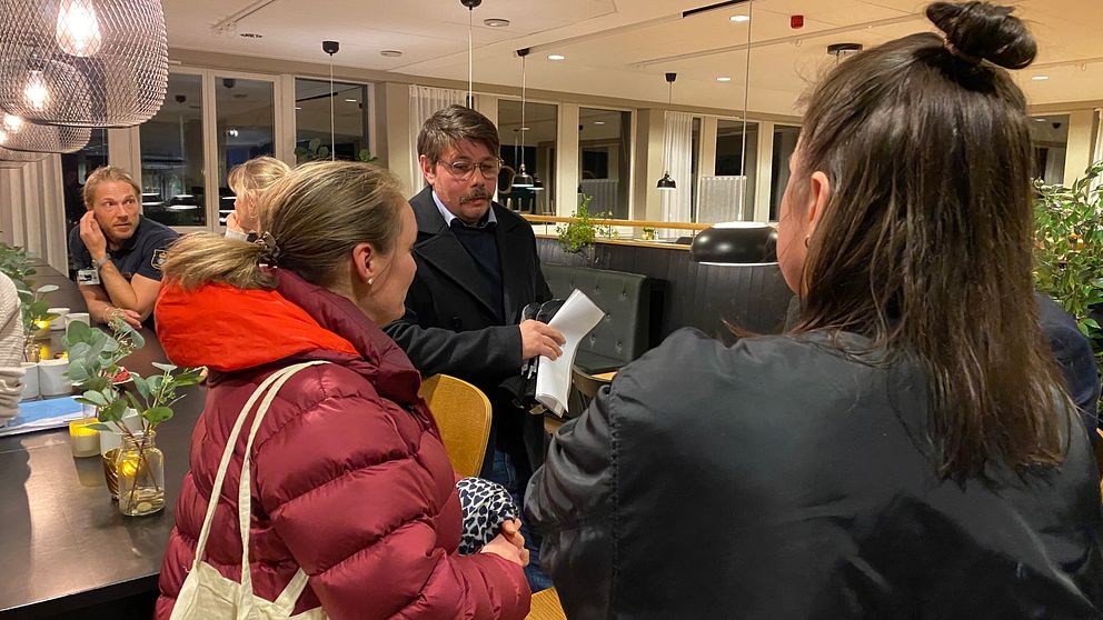 Tre personer samtalar i en inbjudande restaurangmiljö. En kvinna i en röd jacka vänder sig mot en man med dokument i handen, medan en annan kvinna står i bakgrunden. Belysningen i rummet är mjuk och det finns växter på bordet som ger en hemtrevlig atmosfär.