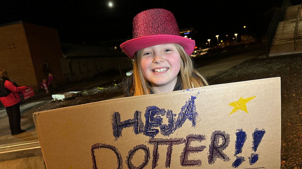 Tjej med glittrig rosa hatt och skylt som tittar in i kameran och ser glad ut