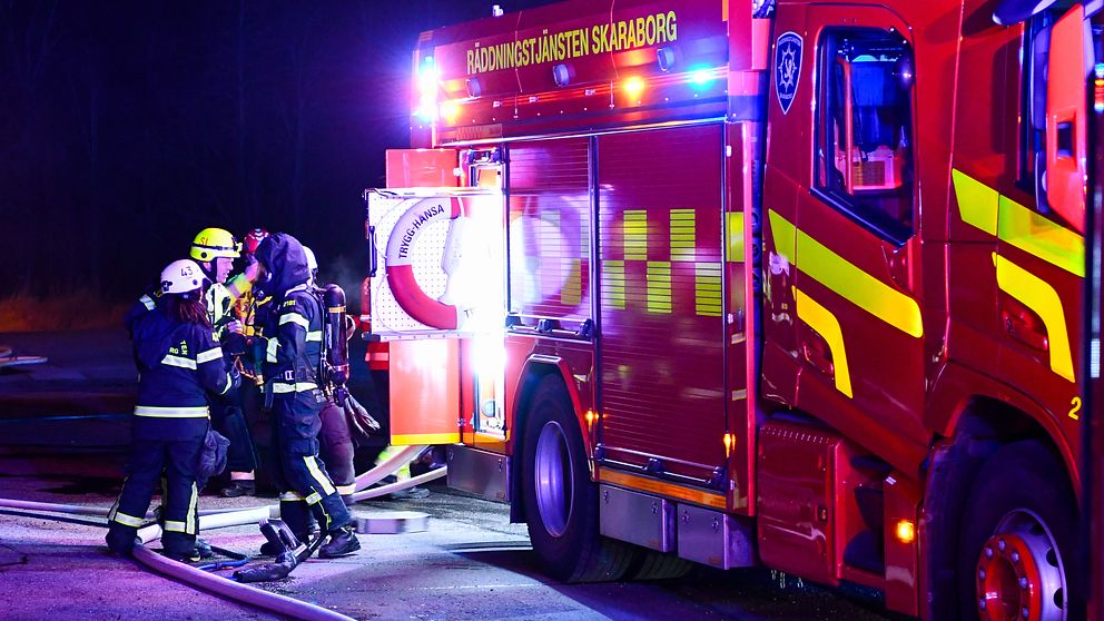 Brandmän i full utrustning arbetar vid en brandbil i mörker, med blå och röda blixtljus som lyser upp scenen. De är engagerade i insatsarbete med släckningsutrustning synlig på marken.