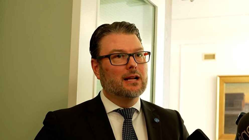 Edward Riedl (M) i kostym intervjuas av flera journalister som håller fram mikrofoner.